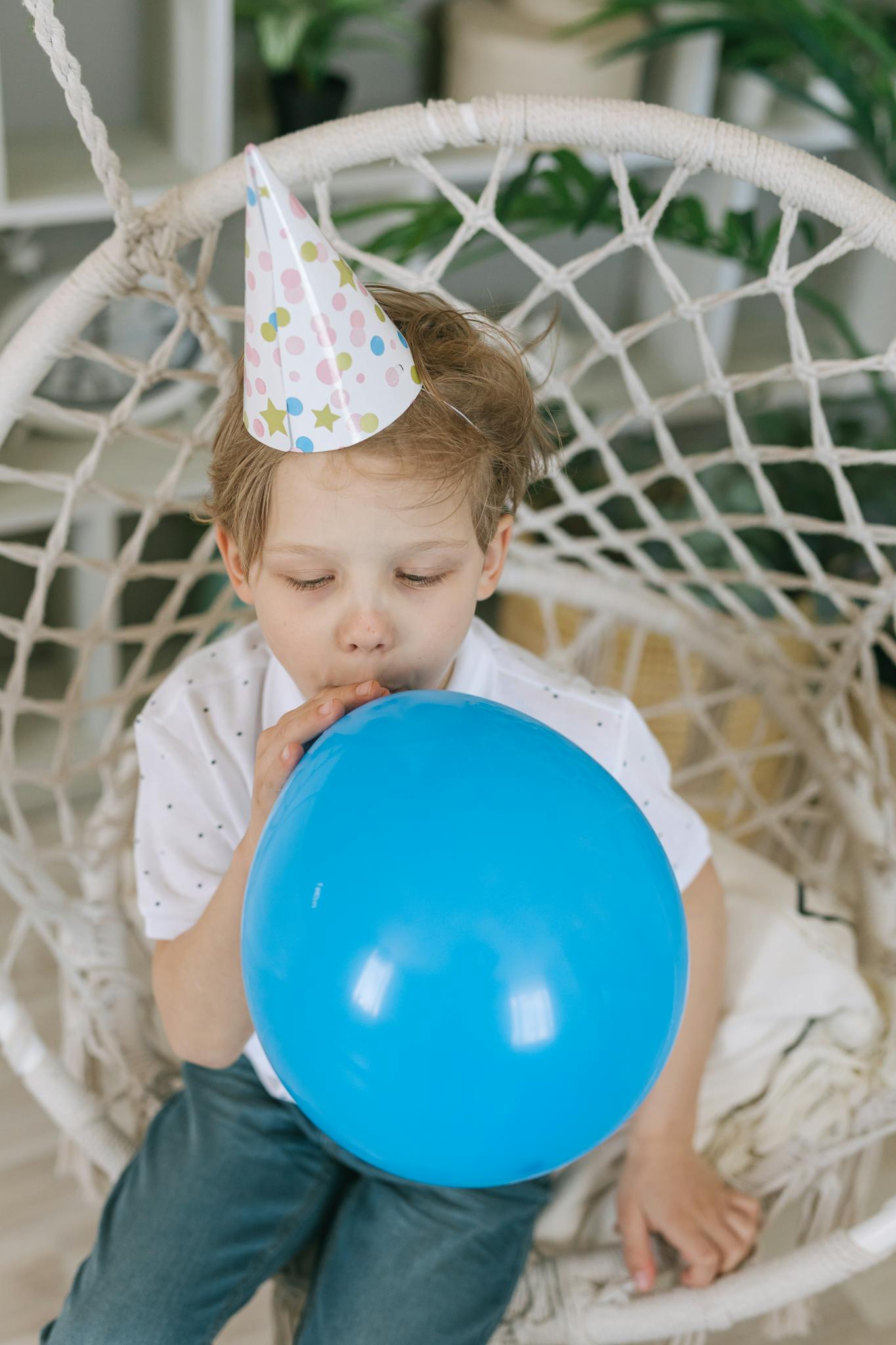 A young boy blowing a blue balloon during a birthday party indoors, wearing a festive hat.