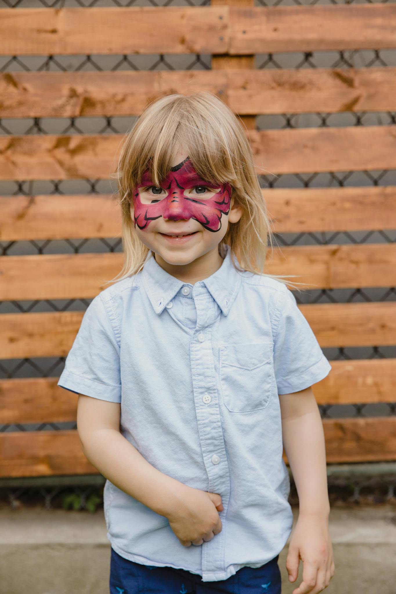 Young child with colorful face paint smiling in front of a wooden fence, enjoying an outdoor activity.